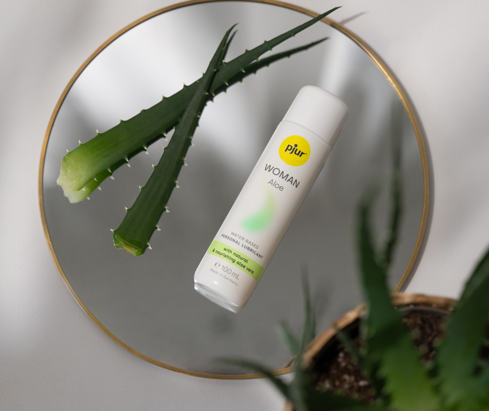 Flasche pjur Woman Aloe auf einem runden Spiegel mit Aloe-Vera-Blättern im Hintergrund.